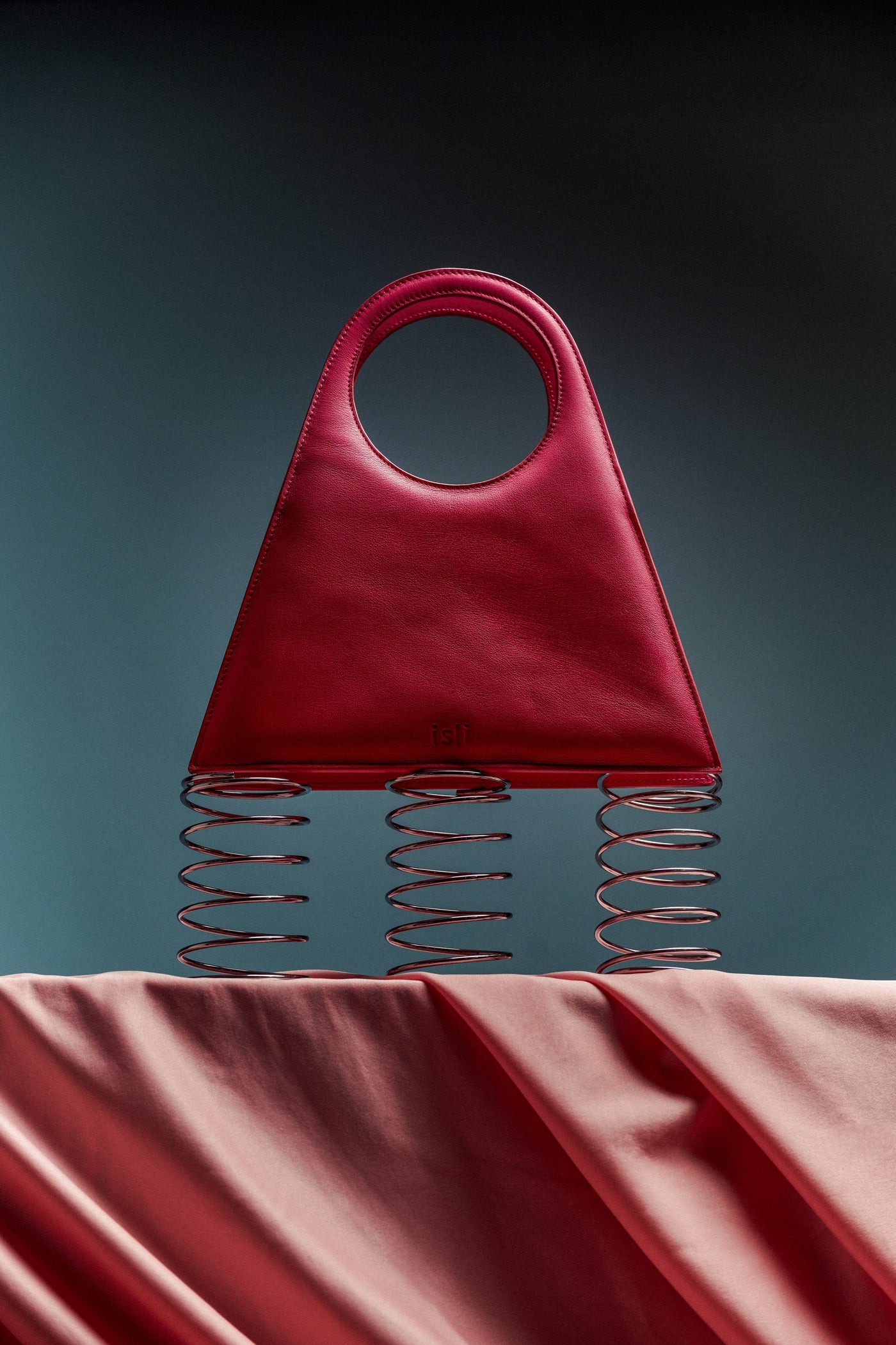 A red ISLI Studio bag with a circular handle displayed on metal springs against a teal backdrop