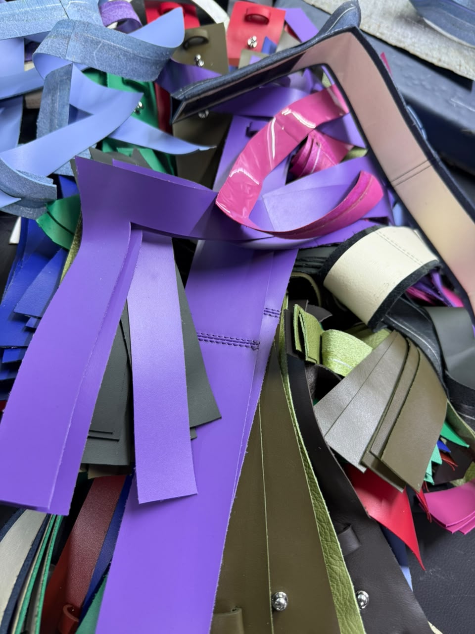 Offcuts of deadstock leather in purple, pink, olive and blue, piled on the workbench.